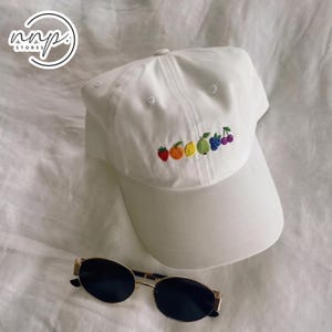 May include: White baseball cap with embroidered colorful fruit design, including a strawberry, orange, lemon, lime, blueberries, and cherries. Gold-rimmed sunglasses with dark lenses are also in the image.