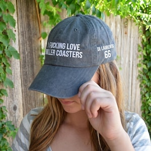 Puede incluir: Una gorra de béisbol gris carbón con el texto blanco I FUCKING LOVE ROLLER COASTERS en el panel frontal y el texto blanco DI LAURENTIS 66 en el panel lateral. La gorra tiene una visera curva y una corona estructurada con ojales cosidos.