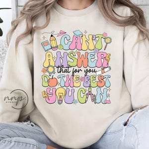 May include: A light beige sweatshirt with a colorful graphic print that says "I can't answer that for you, do the best you can." The graphic includes a pencil, a book, a lightbulb, a compass, a graduation cap, and other school-related items.