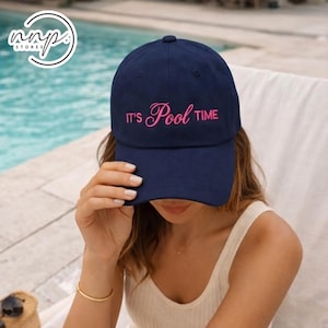 Op de afbeelding: Marineblauwe baseballcap met de roze tekst "IT'S Pool TIME". De cap wordt door een persoon gedragen, met een zwembad op de achtergrond. De cap heeft een gebogen klep en een ronde kroon.