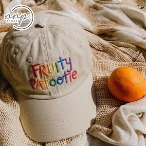 Peut inclure: Casquette de baseball beige avec l'inscription brodée "Fruity Patootie" aux couleurs de l'arc-en-ciel. Un fruit orange est posé à côté de la casquette sur une surface texturée. La casquette a une visière incurvée et un dessus arrondi.