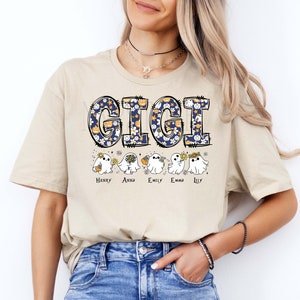 K&ouml;nnte beinhalten: Ein beiges Kurzarm-T-Shirt mit dem Wort GIGI in gro&szlig;en marineblauen Buchstaben mit einem wei&szlig;en Blumen- und K&uuml;rbismuster. Unter dem Text befinden sich f&uuml;nf wei&szlig;e Geisterillustrationen, die jeweils eine Blume oder einen K&uuml;rbis halten, mit den Namen Henry, Anna, Emily, Emma und Lily darunter.