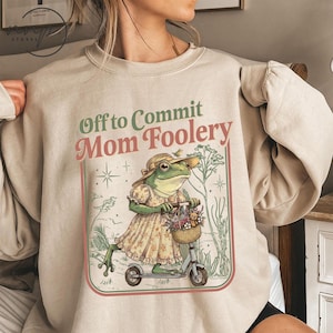 May include: Beige sweatshirt with the text "Off to Commit Mom Foolery." The graphic features a frog in a dress and hat riding a scooter with a basket of flowers and a teddy bear. The design is framed with a red and green border.