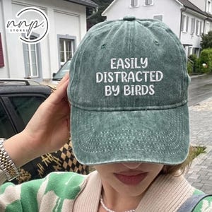 Op de afbeelding: Een groene baseballcap met de tekst "EASILY DISTRACTED BY BIRDS" in het wit geborduurd. De cap is gemaakt van een zacht, versleten materiaal en heeft een gebogen klep. De cap is een casual accessoire.