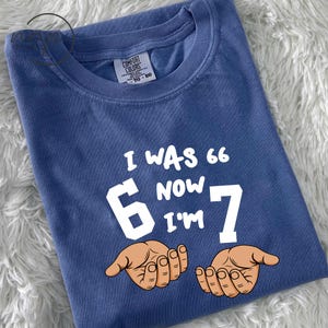 May include: A blue Comfort Colors t-shirt with the text "I WAS 66 NOW I'M 7" in white. The number 6 is in a white block, and the number 7 is in white. Two cartoon hands are below the numbers.