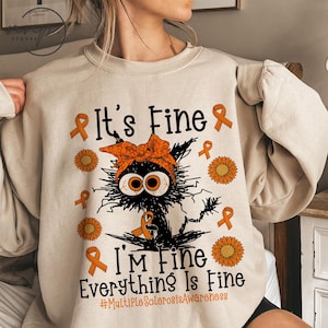 Może przedstawiać: Beżowa bluza z nadrukiem czarnego kota w pomarańczowej bandanie. Pod kotem znajduje się napis "It's Fine, I'm Fine, Everything Is Fine", a także pomarańczowe wstążki i słoneczniki. Dołączono również hashtag #MultipleSclerosisAwareness.
