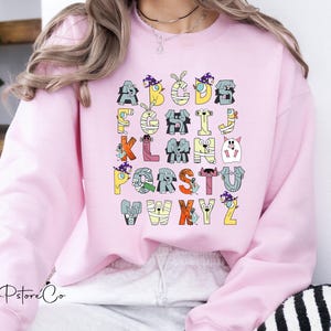 May include: A pale pink sweatshirt displaying a Halloween-themed alphabet. Each letter is designed as a different spooky character, in colours like yellow, orange, grey, and purple. The sweatshirt has a round neck and long sleeves.