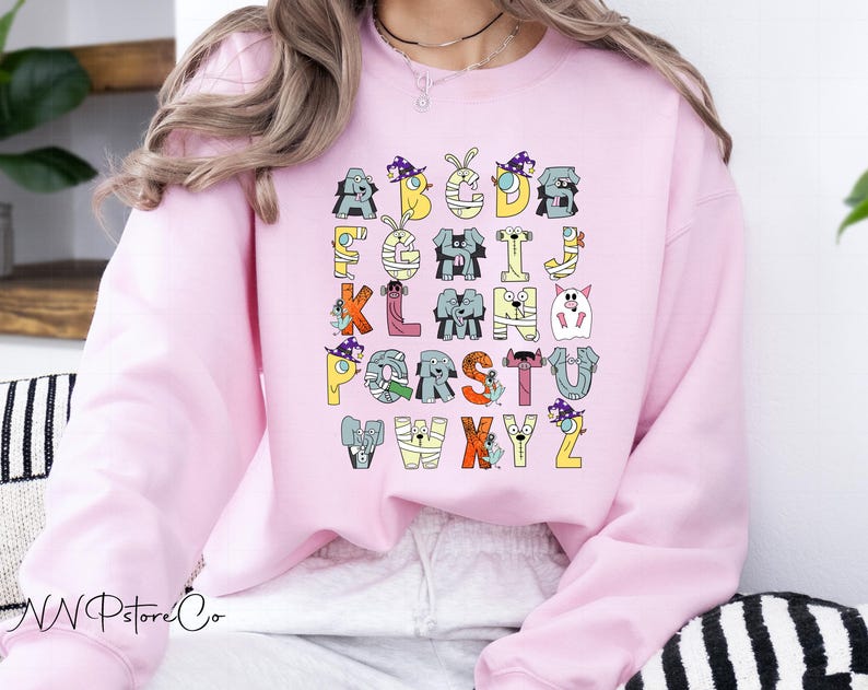 May include: A pale pink sweatshirt displaying a Halloween-themed alphabet. Each letter is designed as a different spooky character, in colours like yellow, orange, grey, and purple. The sweatshirt has a round neck and long sleeves.