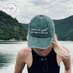 Peut inclure: Casquette de baseball turquoise avec l'inscription blanche "literally just a girl who wants to have fun". La casquette est portée par une personne aux cheveux blonds, avec un lac et des montagnes en arrière-plan.