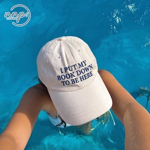 May include: A light beige baseball cap featuring the dark blue embroidered text I PUT MY BOOK DOWN TO BE HERE on the front panel. The cap has a structured crown, a curved brim, and small ventilation eyelets on the top panels of the fabric.