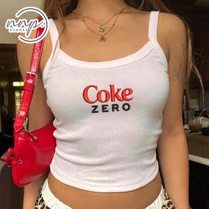 以下が含まれることがあります： 胸元に赤で刺繍されたCokeの文字と、黒で刺繍されたZEROの文字が特徴の白いノースリーブタンクトップ。この衣類はスクープネックと太いショルダーストラップを備えています。鮮やかな赤のショルダーバッグが左側に配置されています。
