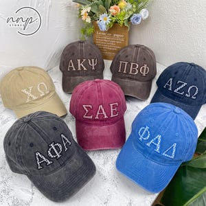May include: A collection of seven baseball caps in various colours including tan, dark grey, brown, maroon, navy blue, and bright blue. Each cap features three-letter Greek-style embroidered patches with a white and blue checkered pattern.