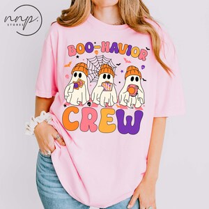 Pode incluir: Camiseta rosa claro com o texto "BOO-HAVIOR CREW" em laranja, rosa e roxo. A camiseta apresenta três fantasmas de desenho animado usando gorros laranja e segurando itens com tema de Halloween. Uma teia de aranha e morcegos também estão incluídos.