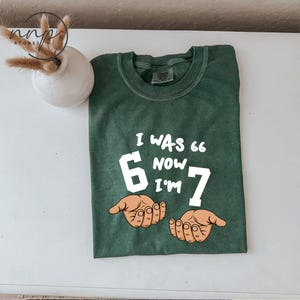 May include: Green t-shirt with the text "I WAS 6 NOW I'M 7" in white. The number 6 is in a white box, and the number 7 is in white. Two hands are drawn below the text. The shirt is on a white surface.