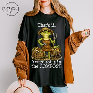 May include: Black t-shirt featuring a frog gardener with the text "That's it. You're going in the COMPOST." The frog wears a straw hat and apron, holding a pitchfork. Wooden crates and a wheelbarrow are filled with vegetables.