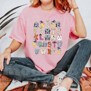 May include: A light pink t-shirt with a Halloween-themed alphabet design. Each letter is decorated with a different character, such as a mummy or a ghost. The letters are in various colours, including yellow, blue, and orange. The shirt is worn by a person.