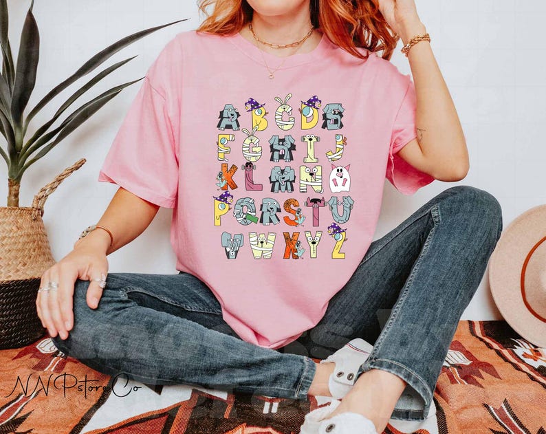 May include: A light pink t-shirt with a Halloween-themed alphabet design. Each letter is decorated with a different character, such as a mummy or a ghost. The letters are in various colours, including yellow, blue, and orange. The shirt is worn by a person.