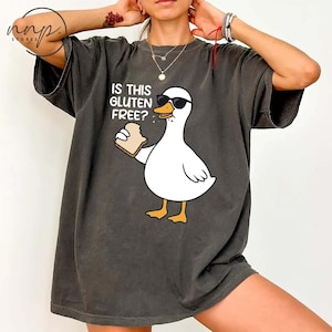 May include: Dark grey t-shirt featuring a cartoon goose wearing sunglasses and holding a piece of bread. The text "IS THIS GLUTEN FREE?" is above the goose. The shirt has a relaxed fit.