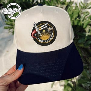 Puede incluir: Gorra de béisbol blanca y azul marino con un parche circular con el texto "PROJECT HAIL MARY". El parche representa un cohete, un satélite y un diseño de estrella. La gorra tiene una visera curva y un botón en la parte superior.