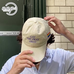 Può includere: Un cappellino da baseball beige con la scritta viola "GAY THE PRAY AWAY". Il cappellino presenta una bandiera arcobaleno sul lato. Il cappellino è un classico design a sei pannelli.