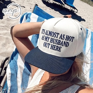 May include: A white baseball cap with a dark blue brim and dark blue embroidered text that reads: IT'S ALMOST AS HOT AS MY HUSBAND OUT HERE. The cap features a structured front panel and a button on the top, designed for sun protection.