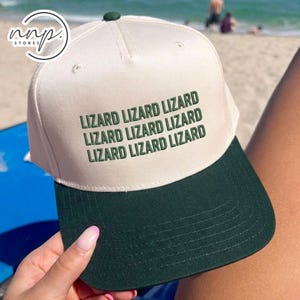 May include: A baseball cap with a cream-colored crown and a dark green brim. The cap features the text "LIZARD LIZARD LIZARD" in green, repeated three times across the front. The cap is being held by a person on a beach.