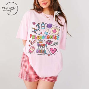 May include: A light pink t-shirt with a colorful graphic that says "Phlebotomist" with medical-themed illustrations. The shirt also has the text "I stab people" and "You give me butterflies".