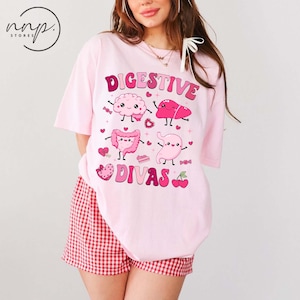 May include: A light pink t-shirt with the words "DIGESTIVE DIVAS" in pink and a cartoon illustration of internal organs. The shirt is paired with red and white checkered shorts. The image includes the logo of "nnp. stores" in a circle.