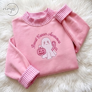 Op de afbeelding: Een lichtroze sweatshirt met een opstaande kraag en mouwboorden in een roze en wit vichy-ruitpatroon. Het midden toont een geborduurd ontwerp van een geest die een pompoen en een roze lint vasthoudt met de tekst Breast Cancer Awareness.