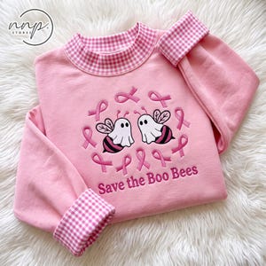 Op de afbeelding: Een lichtroze sweatshirt met een roze en wit gingham kraagje en boorden. Het midden toont een geborduurd ontwerp van twee spookbijen omringd door roze linten en de tekst Save the Boo Bees in roze letters op de voorzijde.