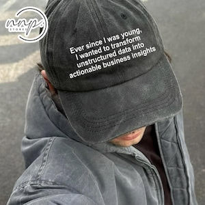 Puede incluir: Gorra de béisbol gris oscuro con el texto blanco "Ever since I was young, I wanted to transform unstructured data into actionable business insights". Llevada por una persona con una chaqueta gris.
