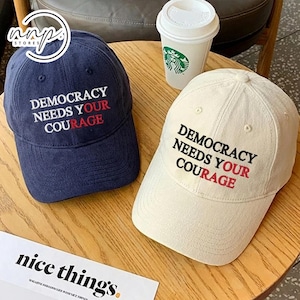Puede incluir: Se muestran dos gorras de béisbol, una azul marino y otra blanquecina. Ambas gorras tienen la frase bordada "DEMOCRACY NEEDS YOUR COURAGE" en blanco y rojo. También se ve un vaso de papel blanco con un logotipo verde.