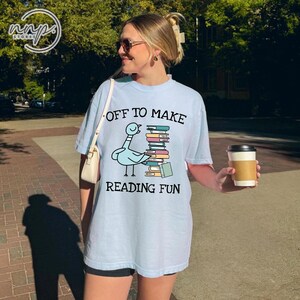 Op de afbeelding: Een lichtblauw t-shirt met korte mouwen en een afbeelding van een blauwe vogel die naast een hoge stapel kleurrijke boeken staat. De tekst "OFF TO MAKE READING FUN" is in het zwart boven en onder de illustratie van de vogel en boeken gedrukt.