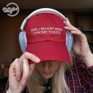 Peut inclure: Une casquette de baseball rouge avec le texte brodé en blanc OOPS, I BOUGHT MORE CONCERT TICKETS sur le panneau avant. La casquette est portée avec un casque audio bleu clair doté d'un arceau blanc et d'accents argentés sur les écouteurs.