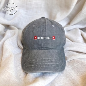 Puede incluir: Una gorra de béisbol gris carbón con acabado desgastado. La parte frontal presenta el texto bordado en blanco DO NOT CALL, flanqueado por dos pequeños iconos rojos que se asemejan a estaciones de alarma contra incendios sobre la visera.
