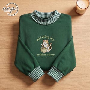 Könnte beinhalten: Ein dunkelgrünes Langarm-Sweatshirt mit einem grün-weiß karierten Kragen und Bündchen. In der Mitte der Brust befindet sich eine Stickerei eines Glückskatzen-Motivs, das in einer Schüssel rührt, mit dem Text whisking my problems away.