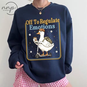 May include: Navy blue sweatshirt featuring a cartoon goose riding a scooter, holding a coffee cup, and wearing a backpack and a brown hat. The text "Off To Regulate Emotions" is displayed in a yellow and blue frame. White daisies and stars are also present.