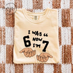 May include: A light yellow t-shirt with the text "I WAS 66 NOW I'M 7" in black. The number 6 is large and bold, with a drawing of a hand on each side. The shirt is made of a soft material.