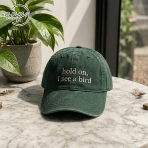Op de afbeelding: Een donker bosgroene baseballpet met de geborduurde tekst "hold on, I see a bird" in lichtbeige draad. De pet heeft een gestructureerde kroon, een gebogen klep en gestikte oogjes op de bovenste panelen voor ventilatie en extra detail.