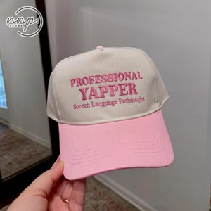 May include: A cream-coloured baseball cap with a pink brim. The front features pink embroidered text that reads PROFESSIONAL YAPPER and Speech Language Pathologist below it. The cap has a structured crown and a curved visor with stitched detailing.