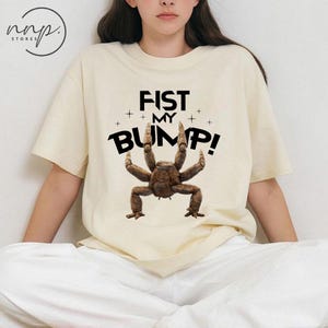 May include: Cream-colored t-shirt featuring the text "FIST MY BUMP!" in black, with a brown crab-like creature graphic. The shirt has short sleeves and a crew neck.