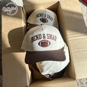 May include: A cream-colored baseball cap with a brown brim featuring the embroidered text BEND & SNAP in brown letters above a brown football graphic. The cap is positioned inside a cardboard box with another identical cap visible behind it.
