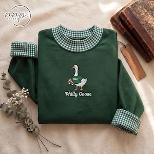 May include: A dark green long-sleeve sweatshirt featuring a white and green gingham collar and sleeve cuffs. The centre displays an embroidered goose wearing a green jersey with the word PHILLY and holding a football, with the text Philly Goose below.