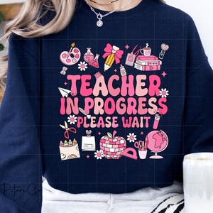 Może przedstawiać: Granatowa bluza z nadrukiem w kolorze różowym i białym, na którym widnieje napis "Teacher in Progress Please Wait". Grafika zawiera różne artykuły szkolne, takie jak ołówek, linijka, globus i książka.