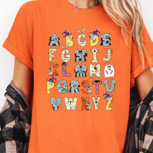 May include: An orange t-shirt with a colourful alphabet design. Each letter is represented by a whimsical animal character in Halloween costumes. The letters are in various colours. The t-shirt has short sleeves.
