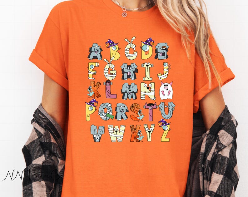 May include: An orange t-shirt with a colourful alphabet design. Each letter is represented by a whimsical animal character in Halloween costumes. The letters are in various colours. The t-shirt has short sleeves.