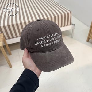 May include: A charcoal gray baseball cap featuring white embroidered text that reads, I THINK A LOT OF MY PROBLEMS WOULD BE SOLVED IF I HAD A DRAGON. The cap has a structured crown, a curved brim, and stitched eyelets on the top panels.