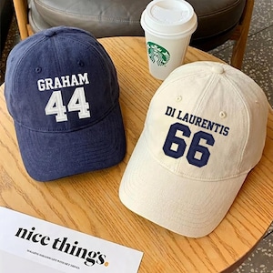 May include: Two baseball caps, one navy blue and one off-white, sit on a wooden table. The navy cap has "GRAHAM" and the number "44" embroidered in white. The off-white cap features "DI LAURENTIS" and the number "66" in navy. A coffee cup is visible.