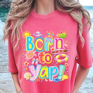 May include: Pink t-shirt with a colourful graphic that reads "Born to Yap!" The design includes a sun, hat, sunglasses, coconut, turtle, and other beach-themed elements. The shirt is a comfortable, casual style.