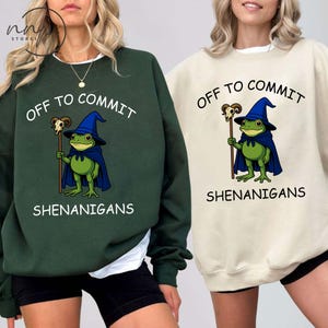 以下が含まれることがあります： 2つのスウェットシャツ。1つは濃い緑色、もう1つはクリーム色で、それぞれに「OFF TO COMMIT SHENANIGANS」というフレーズが書かれています。デザインは、魔法使いの帽子とマントを着て杖を持った緑色のカエルが特徴です。カジュアルウェアです。